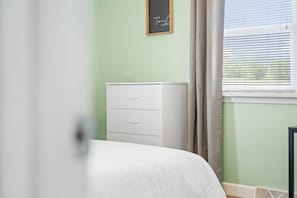 Ein Schlafzimmer mit einem weißen Bett, einer weißen Kommode, einem Fenster mit Jalousien und einer grünen Wand.