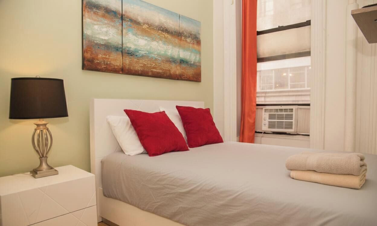 A bedroom with a bed, a nightstand with a lamp, a painting on the wall, and a window with curtains.