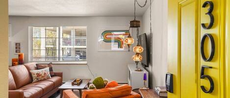 A living room with a sofa, coffee table, and a TV. There is a window with blinds, a lamp, and a door with a number.