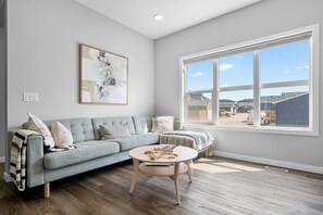 A living room with a light blue sofa, a round coffee table, and a large window with a view of a residential area. - Relax in Comfort with AC/Airport/Stampede (Calgary)