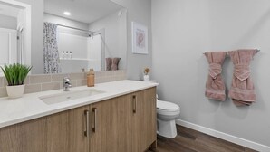 Ein modernes Badezimmer mit einem großen Spiegel, einem Waschbecken und einer Toilette. Handtücher hängen an der Wand und auf der Ablage steht ein Blumentopf.