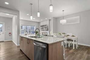 A modern kitchen with a central island, built-in appliances, pendant lighting, and a dining area with white chairs. - Relax in Comfort with AC/Airport/Stampede (Calgary)