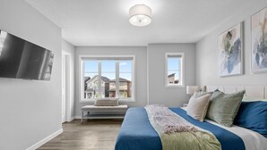 Ein Schlafzimmer mit Bett, einer Bank, einem Fernseher und einem Fenster mit Blick auf Häuser.