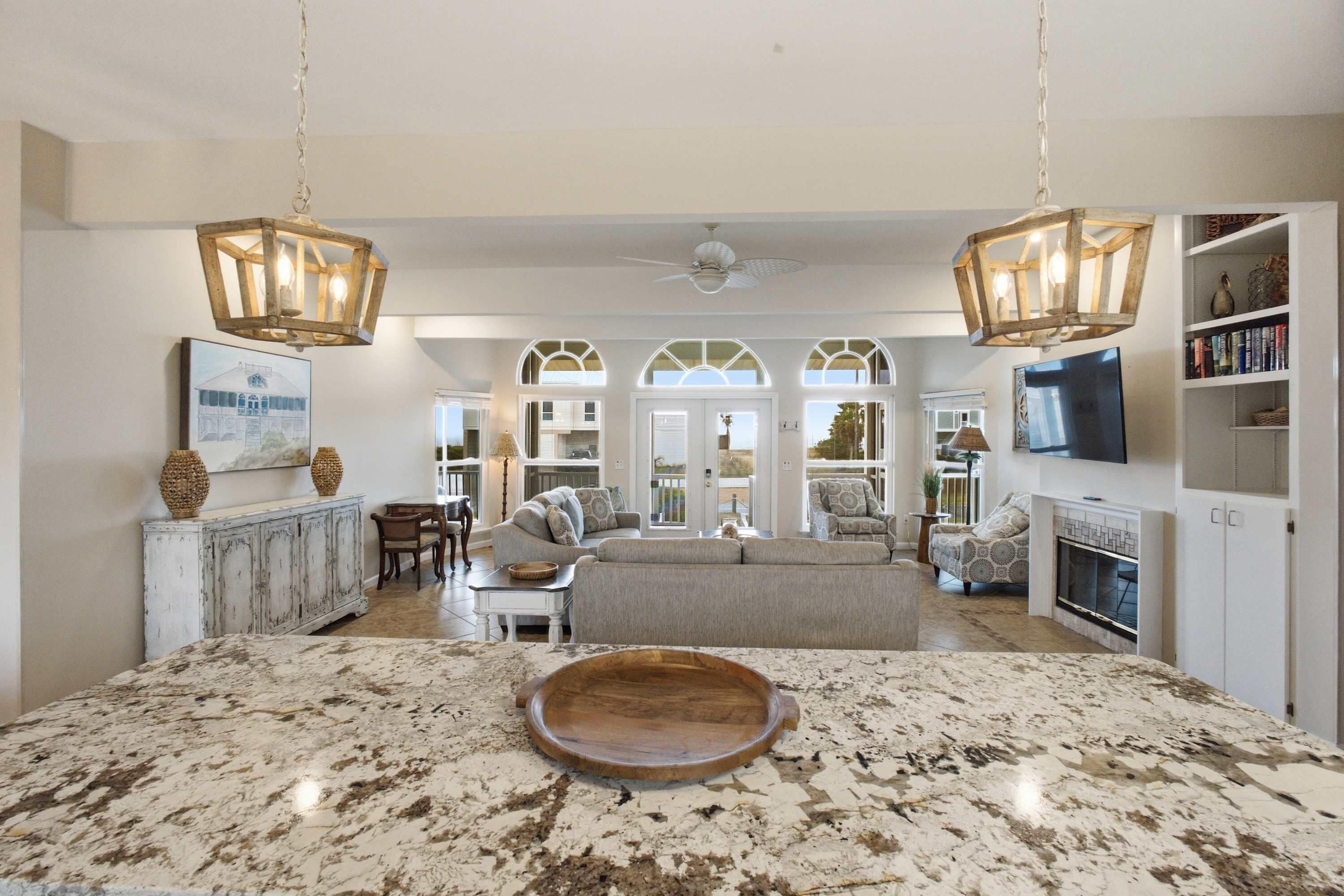 A spacious living area with a large marble island, two wooden chandeliers, a fireplace, and a view through the open doors.