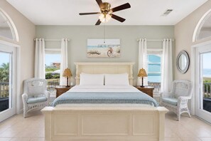 A bedroom with a large bed, two bedside tables, two wicker chairs, a ceiling fan, and a painting on the wall. - Perfect SGI Paradise- 5 Bedroom+ POOL SGI (Saint George Island)