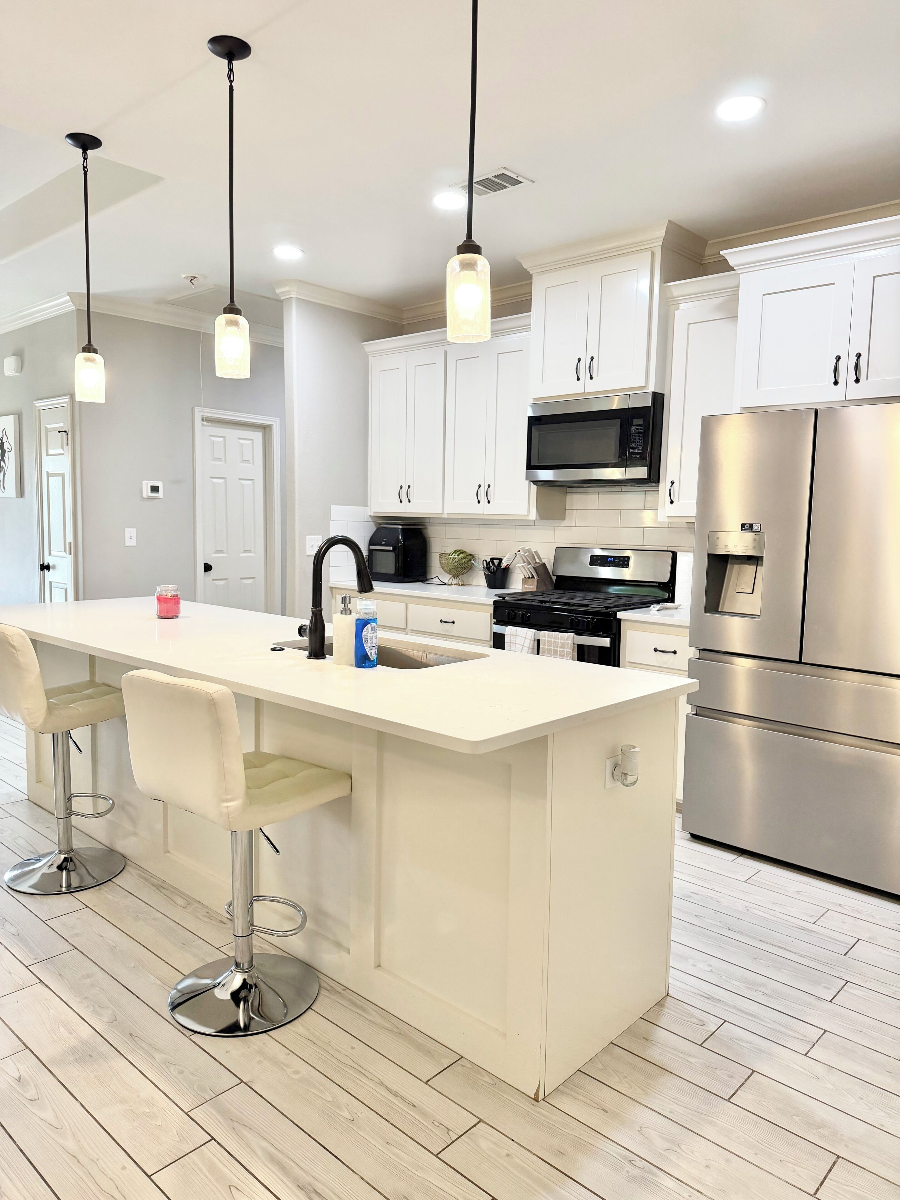 A modern kitchen with white cabinetry, stainless steel appliances, a central island with bar stools, pendant lighting, and a tiled floor.