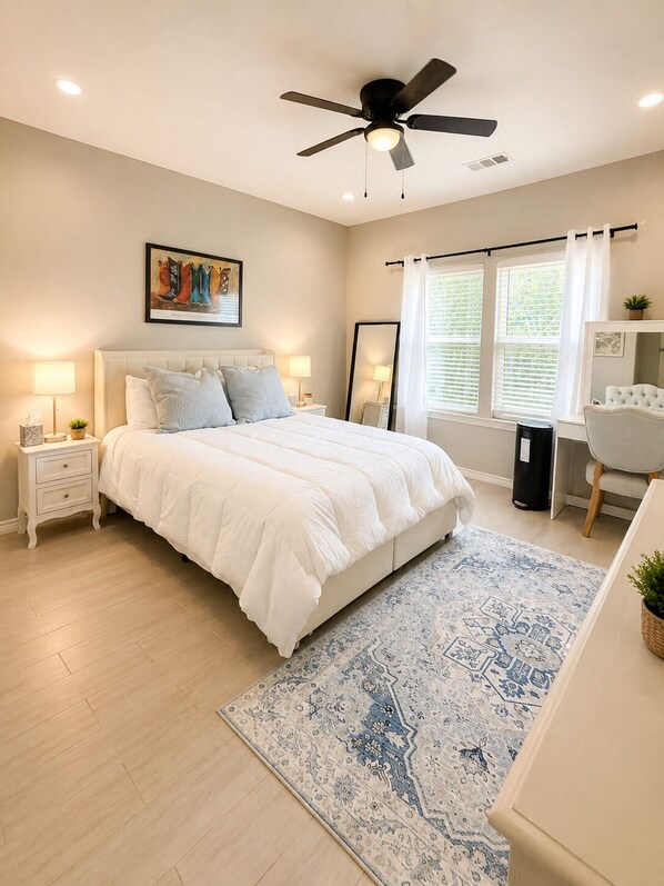 A bedroom with a bed, bedside tables, a ceiling fan, a window with curtains, a mirror, a desk with a chair, and a patterned rug.