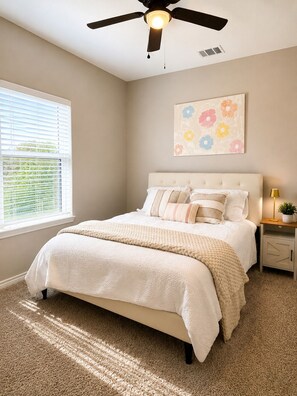 A bedroom with a bed, a ceiling fan, a window with blinds, a nightstand with a lamp, and a wall art piece.