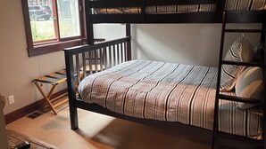 Uma cama beliche com colchão listrado, escada de madeira e janela com vista para um carro estacionado lá fora.
