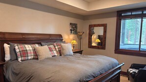 Quarto com uma cama grande, mesa de cabeceira com luminária, espelho e janela com vista para árvores.