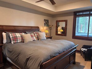 Een slaapkamer met een groot bed, een nachtkastje met een lamp, een spiegel en een raam met uitzicht op bomen.