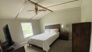 A bedroom with a bed, a nightstand, a desk with a computer, a ceiling fan, and a window with blinds.