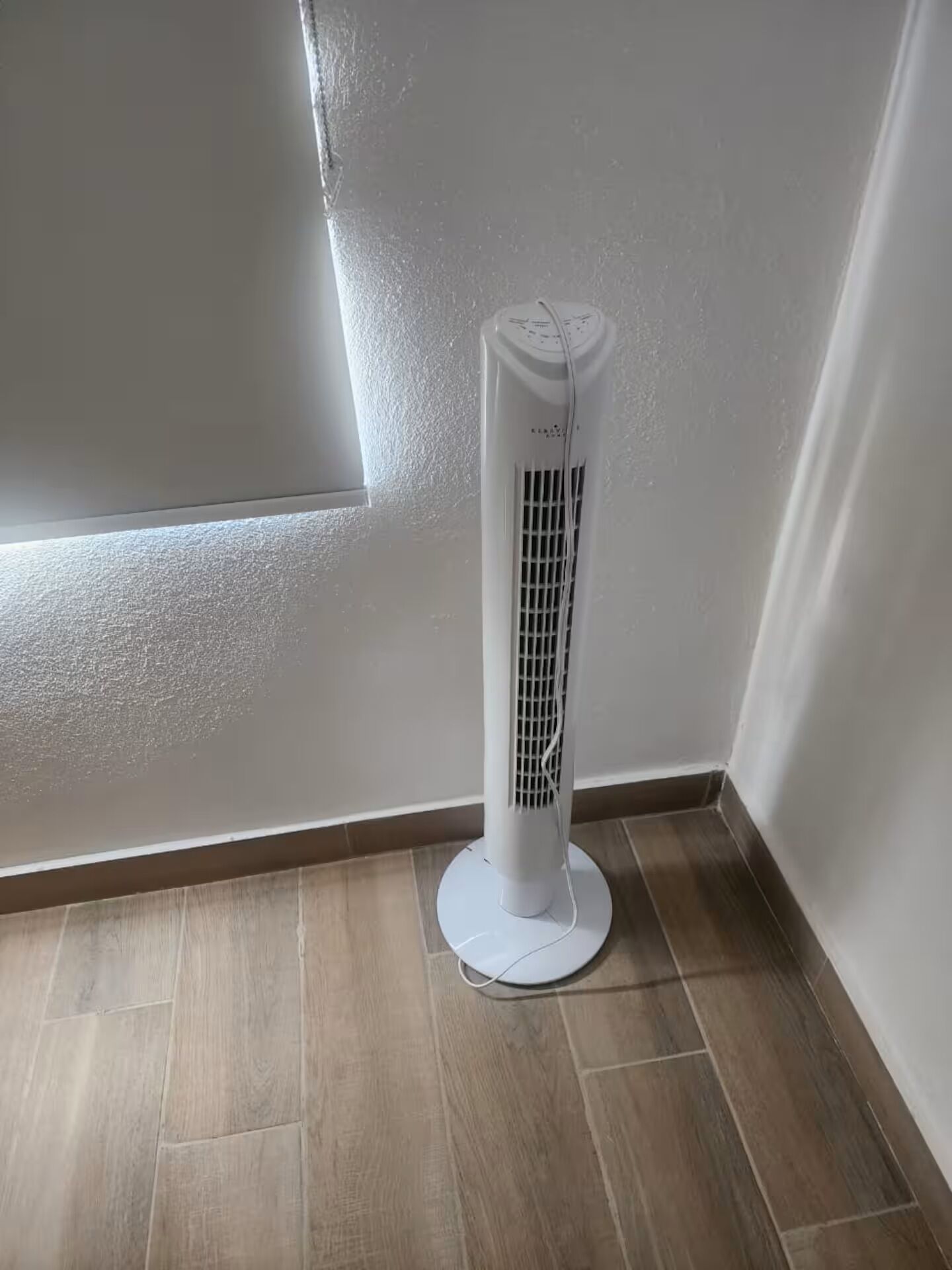 Un ventilateur blanc, posé sur pied, se trouve dans un coin d’une pièce dont le sol est en bois et les murs sont blancs.