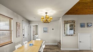 Un coin repas avec une table en bois et des chaises blanches, un lustre et un miroir doté d’un cadre décoratif.