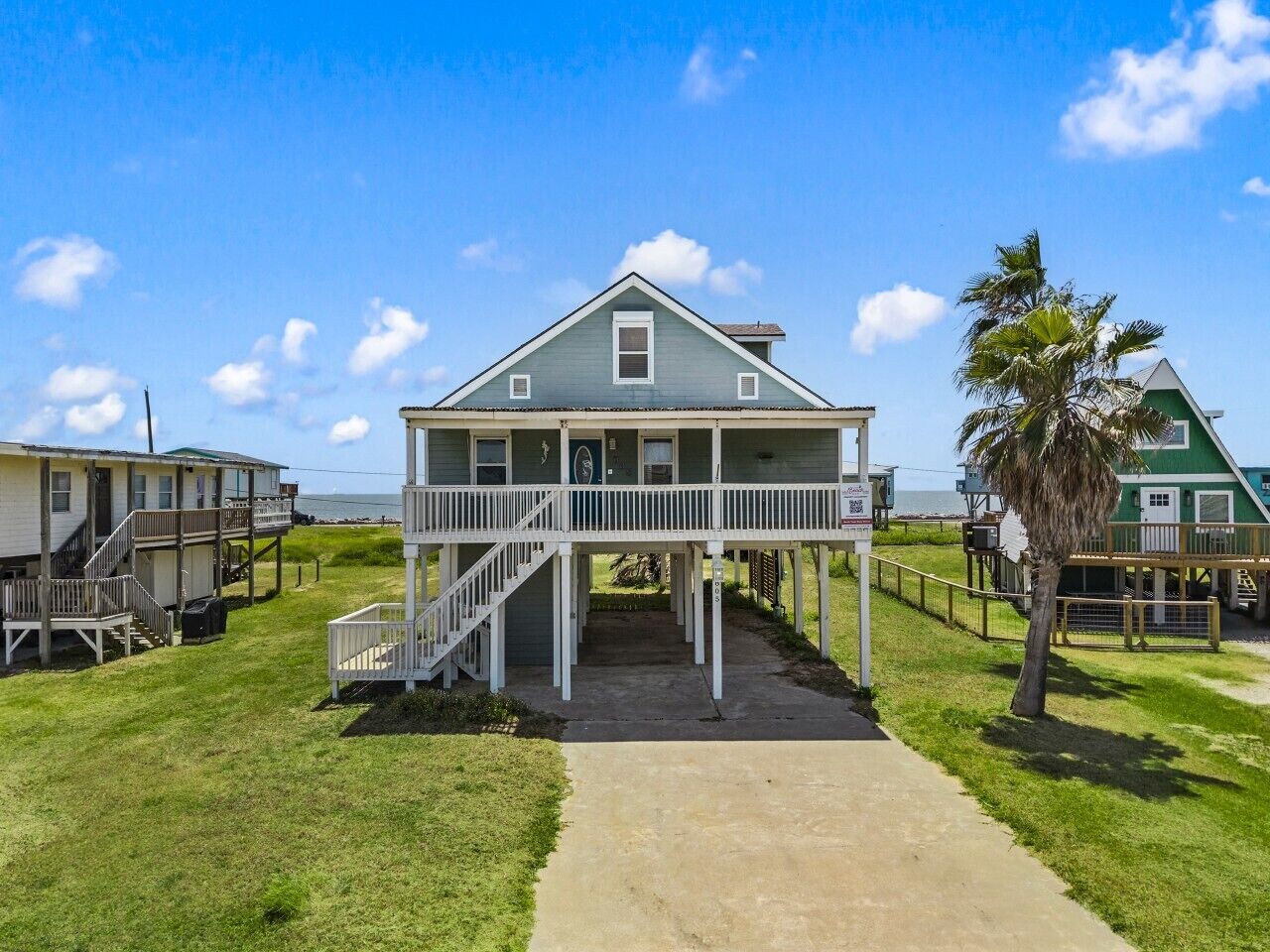 A beachfront property with a central building featuring a porch and stairs, surrounded by a pathway and palm trees.