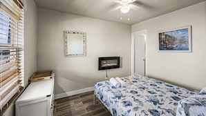 Une chambre à coucher avec un lit, un ventilateur de plafond, une télévision, un tableau et une fenêtre avec des stores.