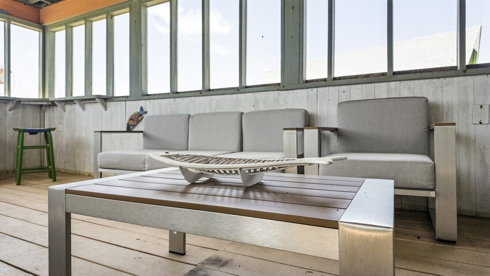 Une véranda avec un plancher en bois, un canapé gris et une table en métal surmontée d’un plateau.