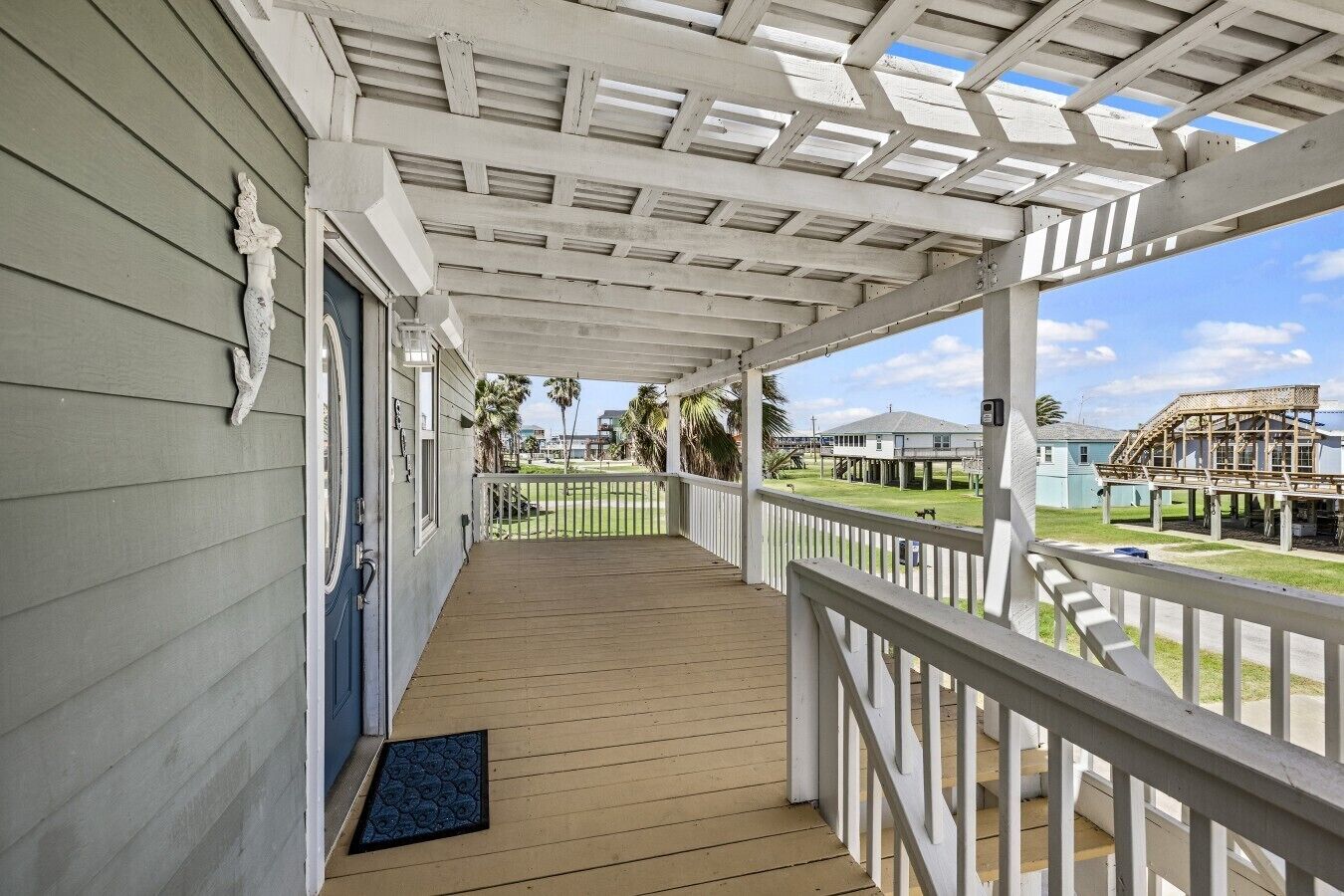 A covered porch with a wooden floor and white railings, overlooking a grassy area with palm trees and a building under construction.