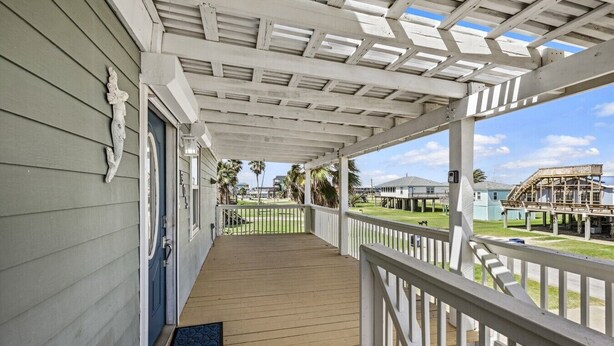 Une véranda couverte, dotée d’un plancher en bois et de balustrades blanches, qui donne sur une pelouse où se trouvent des palmiers et un bâtiment en construction.