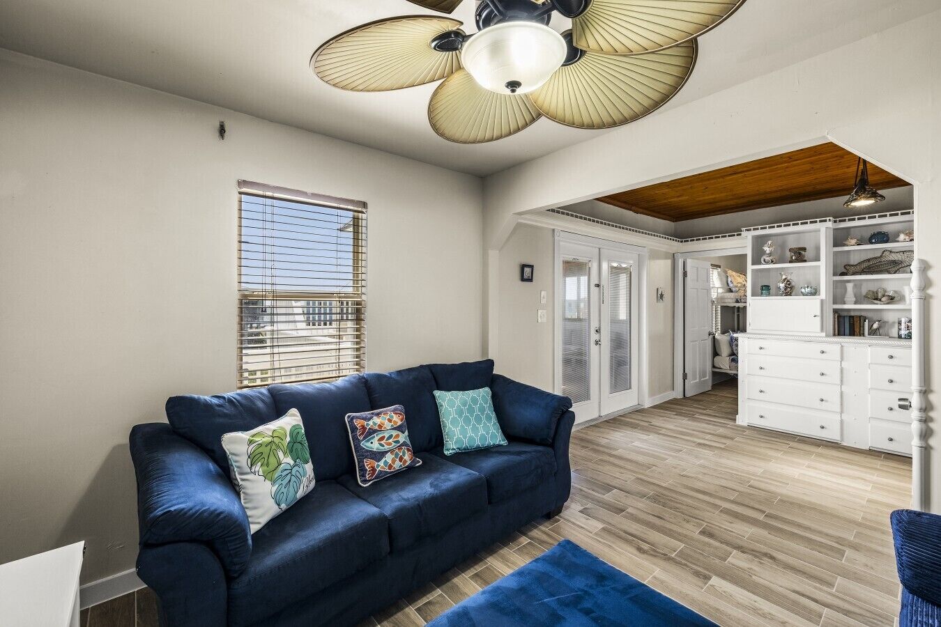 A room with a blue sofa, a ceiling fan, a window with blinds, and a white cabinet with drawers.