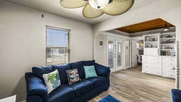 A room with a blue sofa, a ceiling fan, a window with blinds, and a white cabinet with drawers.