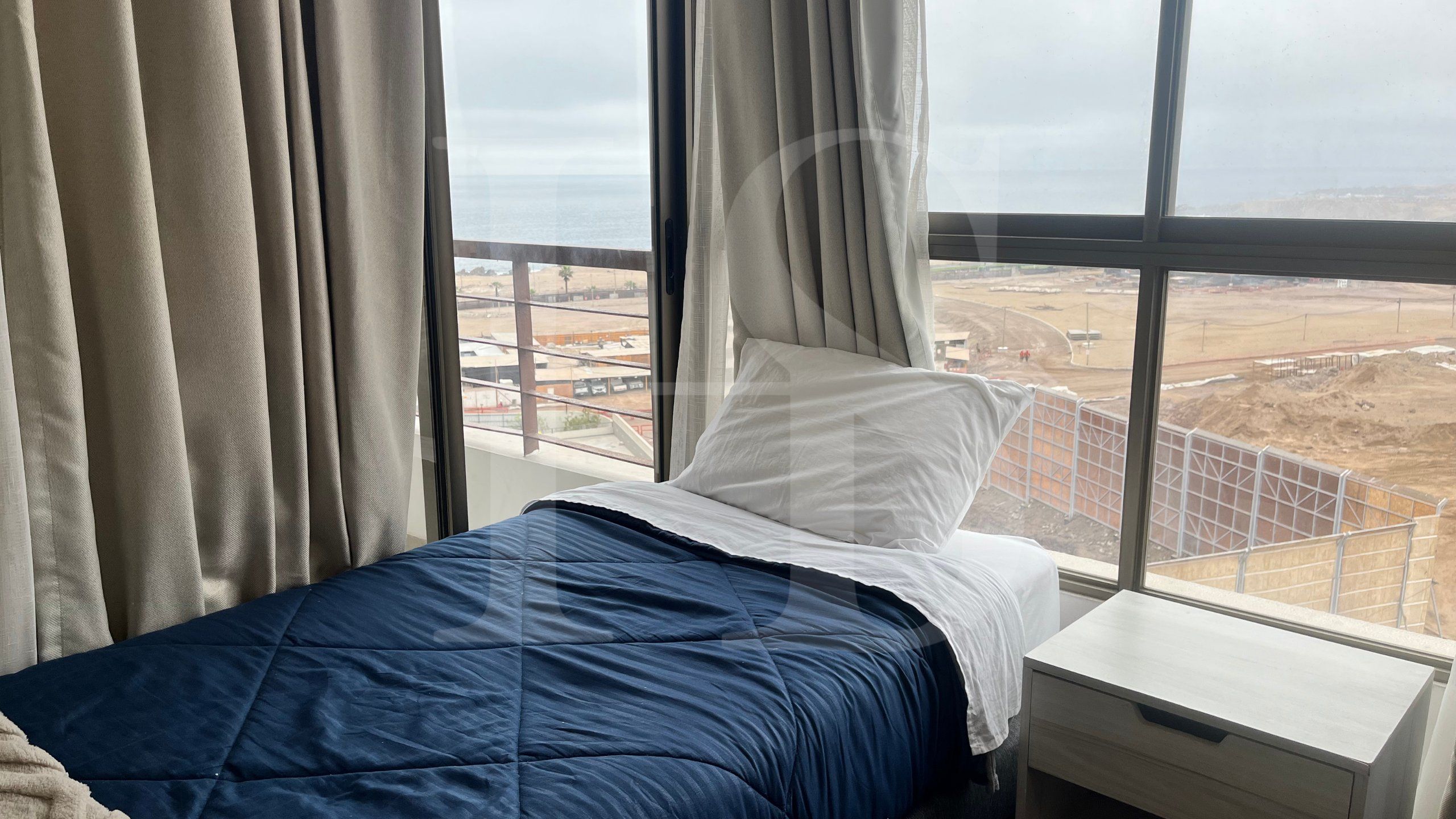 Una cama con colcha azul, almohada blanca y vista a una obra en construcción a través de una ventana con cortinas.