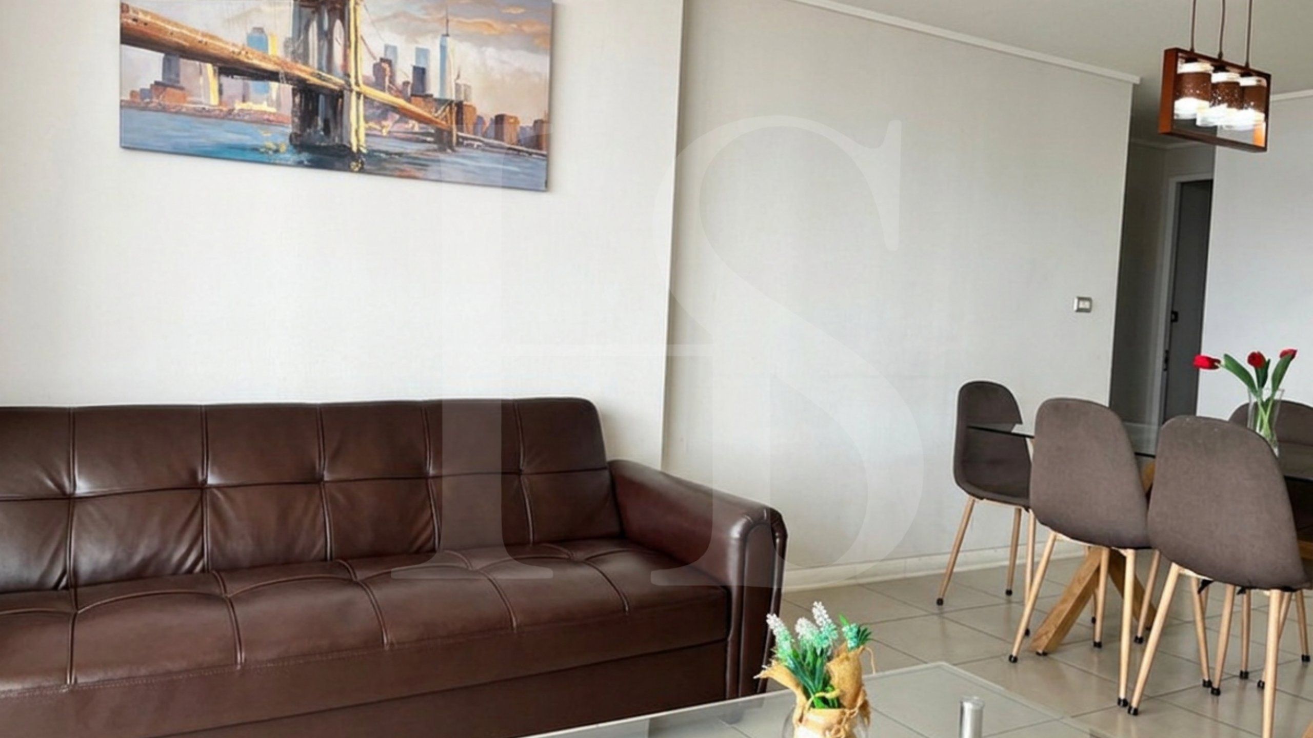 A living room with a brown leather sofa, a dining table with chairs, and a painting of a bridge on the wall.