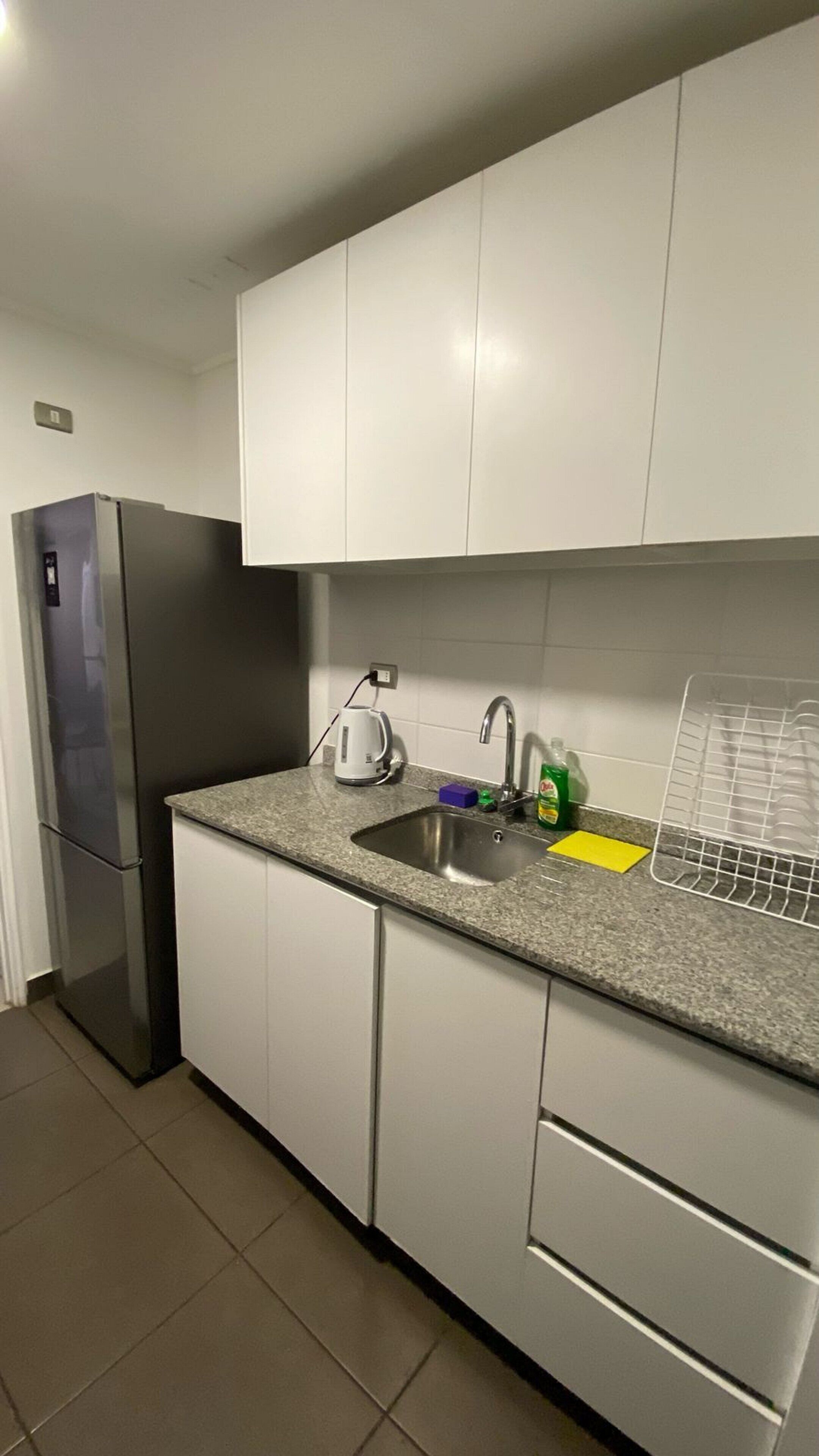 Una cocina con armarios blancos, un refrigerador de acero inoxidable, un lavabo con grifo, una tetera y un escurreplatos.