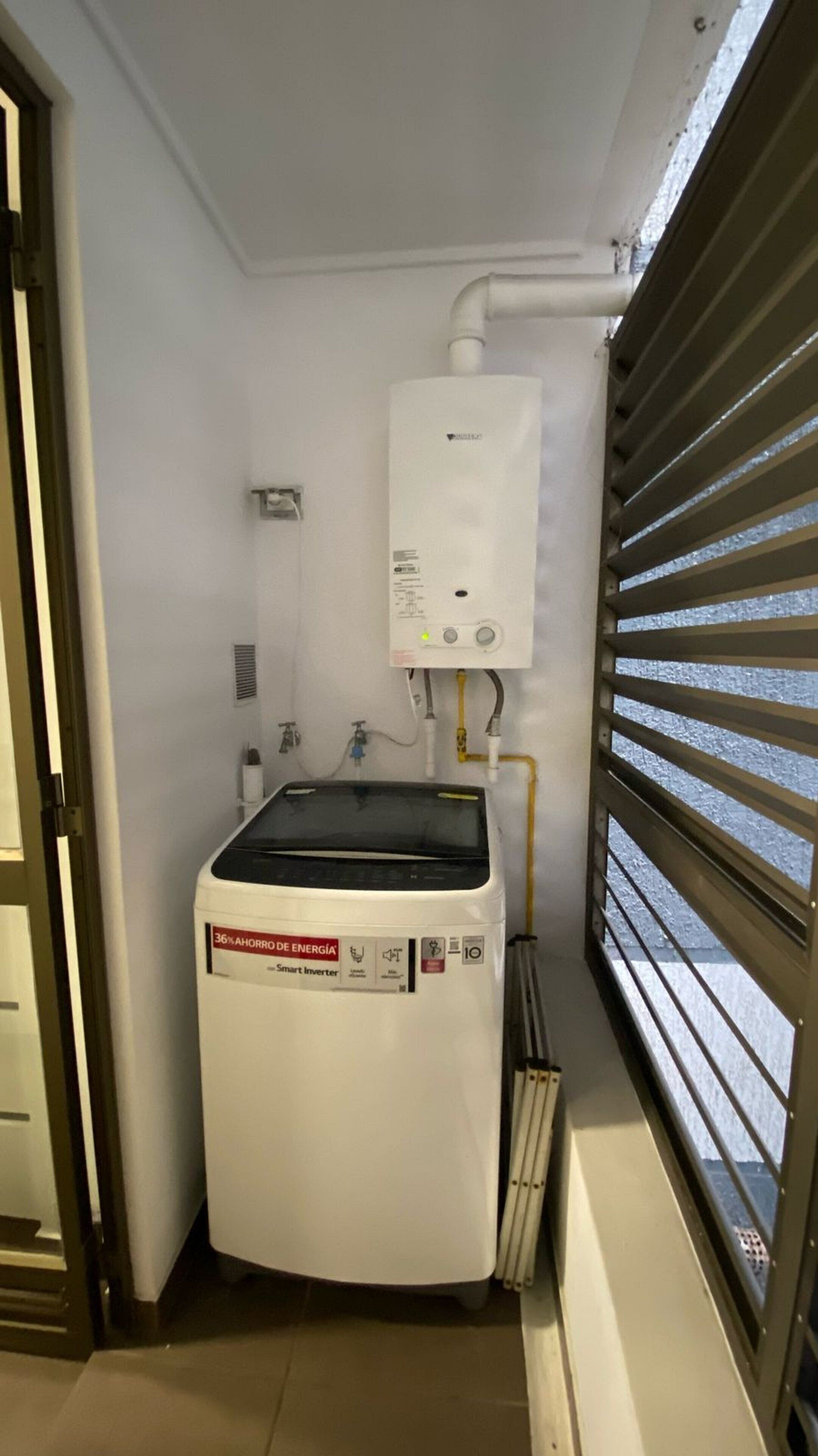Habitación compacta con calentador de agua blanco, radiador blanco y una ventana con persianas horizontales.