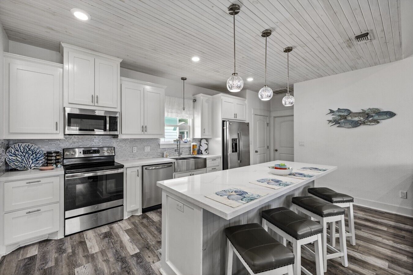 Una cocina moderna con armarios blancos, electrodomésticos de acero inoxidable, una isla central con taburetes y luminarias colgantes.