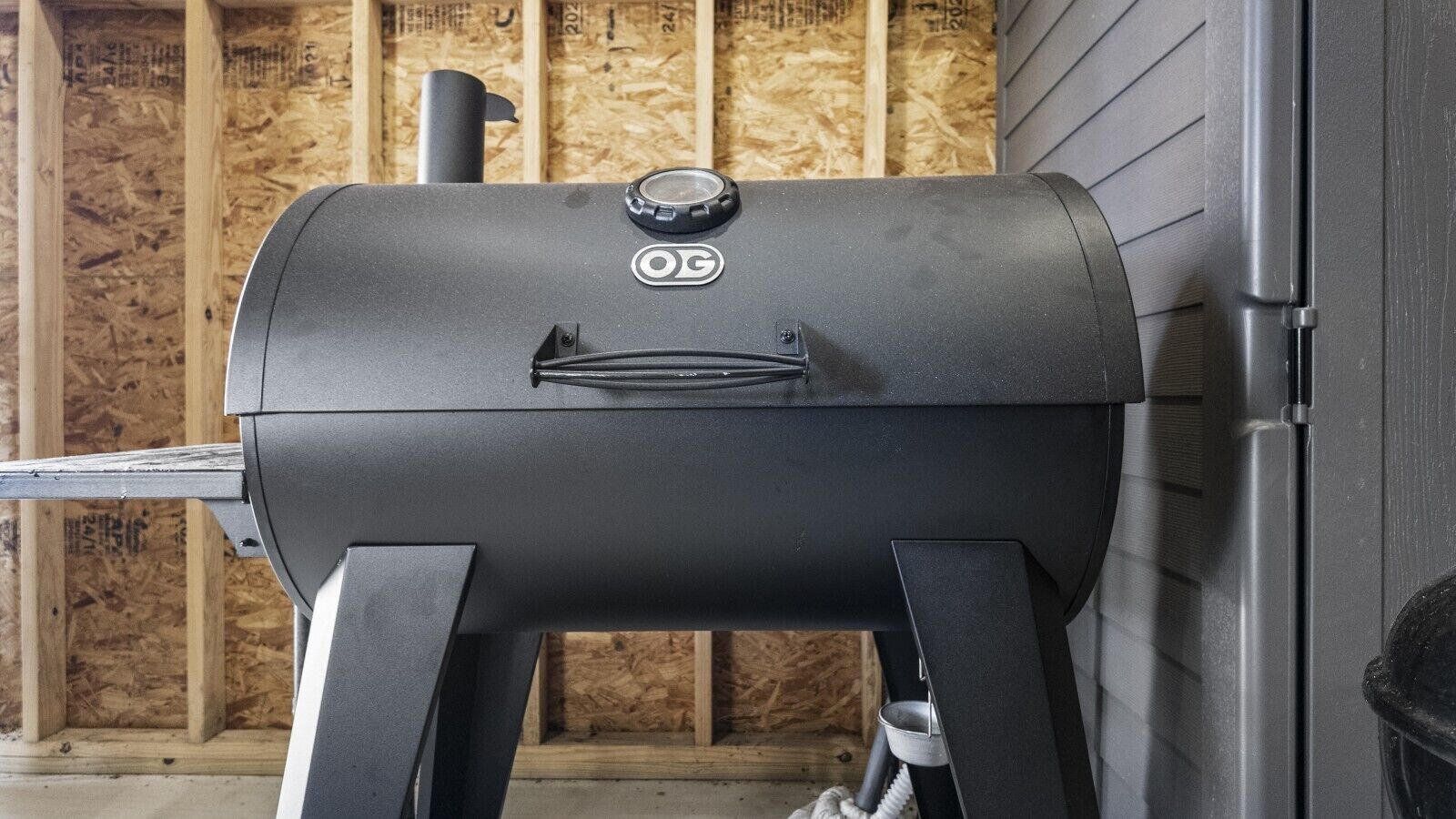 A black outdoor grill with a control knob and a logo, positioned against a wooden wall with insulation.