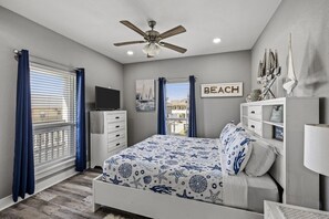 A bedroom with a bed, a dresser, a ceiling fan, a window with curtains, and a wall-mounted TV. - Seadriftin Away - Walk to Beach / Sleeps 12 (Crystal Beach)