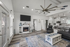 A modern living room with a fireplace, a ceiling fan, and a kitchen area. - Seadriftin Away - Walk to Beach / Sleeps 12 (Crystal Beach)
