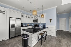 A modern kitchen with white cabinetry, a stainless steel refrigerator, a black countertop island with stools, and a cross on the wall. - Regal Gal | Bird Watcher`s Coastal Escape (Galveston)