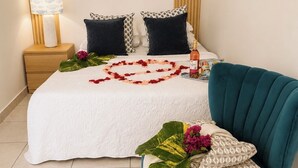 Cama bem arrumada com um arranjo de flores em formato de coração, uma garrafa de champanhe e uma poltrona azul-esverdeada.