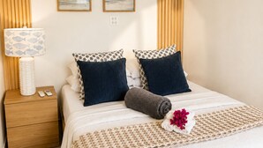 Cama bem arrumada com travesseiro de padrão branco e azul, um travesseiro azul escuro e uma toalha enrolada. Há uma pequena mesa de cabeceira com uma luminária e um telefone.