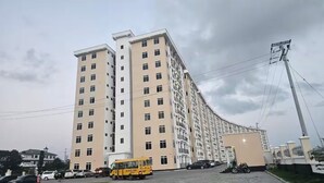 Un edificio residencial de varios pisos con fachada curva, una estructura pequeña adyacente y un vehículo amarillo estacionado.