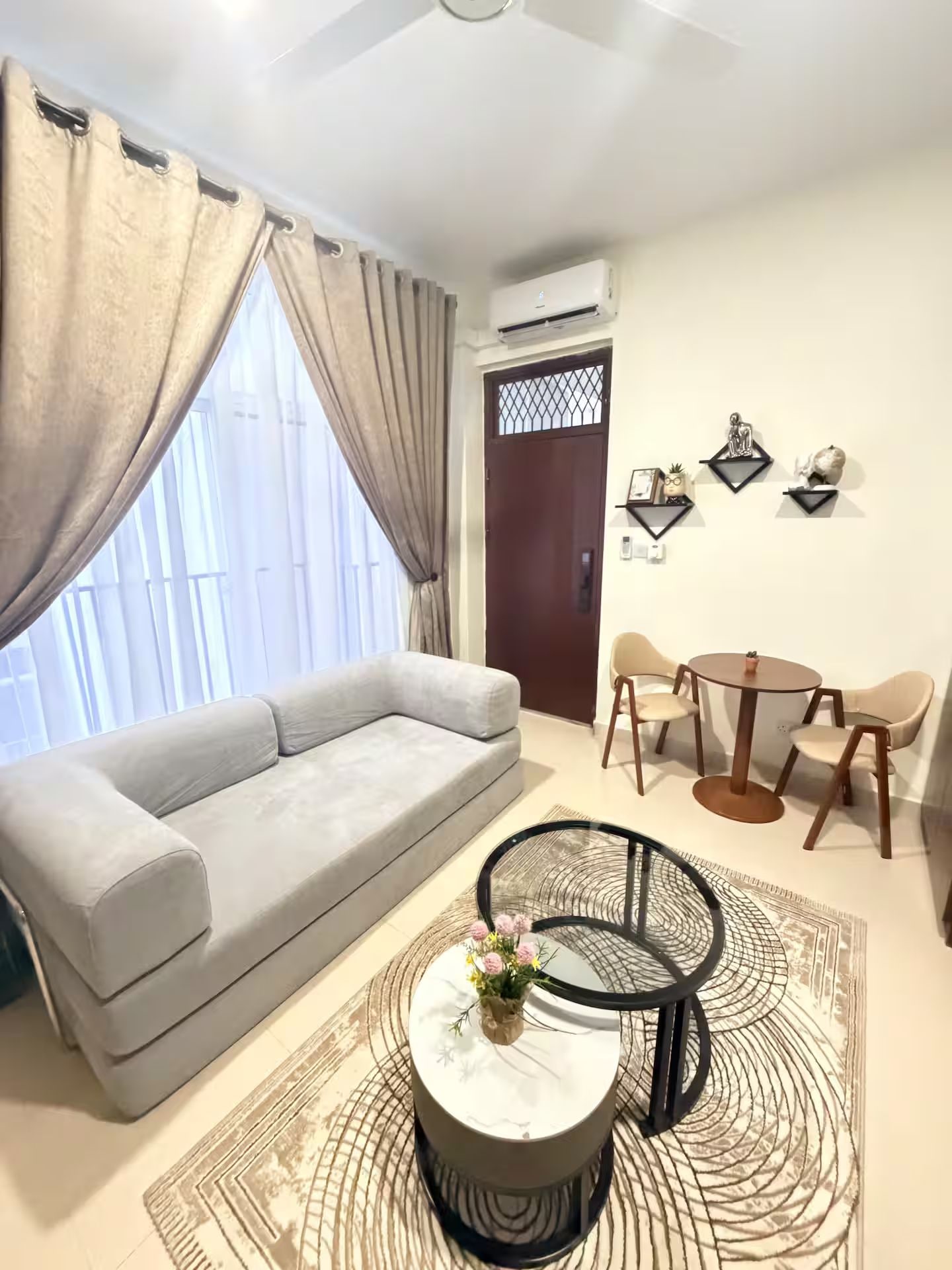 Uma sala de estar com sofá, mesa de centro com vaso, mesa de jantar com cadeiras e um objeto decorativo na parede.