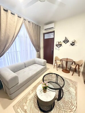 Uma sala de estar com sofá, mesa de centro com vaso, mesa de jantar com cadeiras e um objeto decorativo na parede.