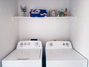 A laundry room with two front-loading washers and a shelf above containing a potted plant, folded towels, and cleaning supplies. - Soleil House - Unit A- Close to beach/downtown (Galveston)