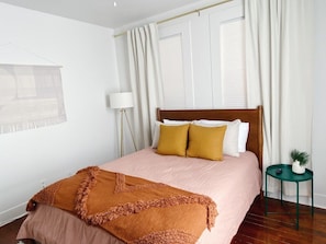 Ein ordentlich gemachtes Bett mit einer rosa Tagesdecke und zwei Kissen, ein kleiner, grüner Nachttisch mit einer Pflanze und eine Stehlampe neben dem Bett.