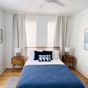 Een slaapkamer met een bed, twee nachtkastjes, een plafondventilator en een raam met gordijnen.