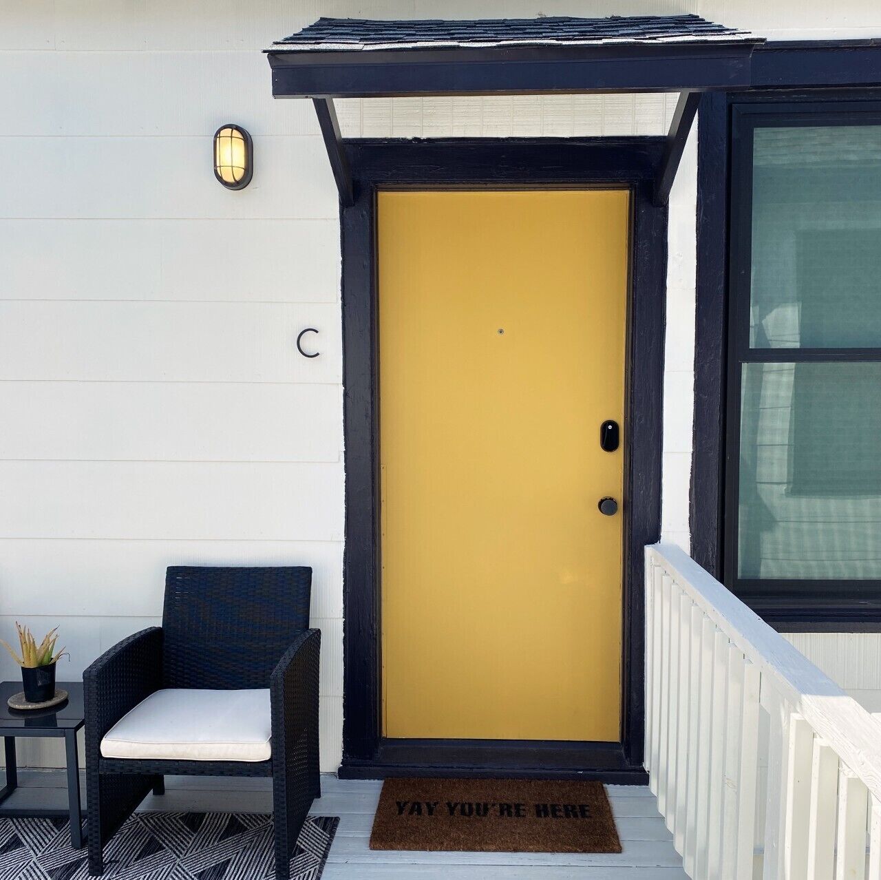A yellow door with a black frame, a small black chair with a white cushion, and a doormat with the text 'YAY YOU'RE HERE'.
