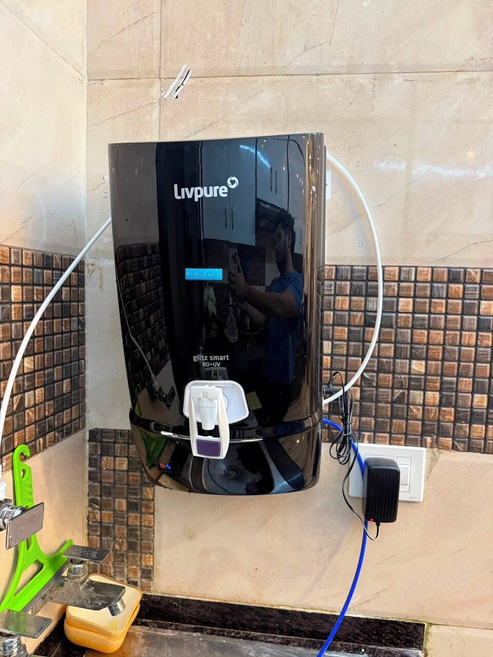 A black Livpure water dispenser mounted on a tiled wall with a visible power cord and a green cleaning tool on the floor.