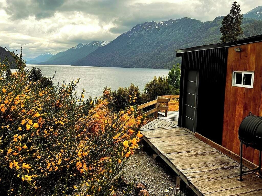 A cabin with a wooden deck overlooking a lake, surrounded by mountains and autumn foliage.