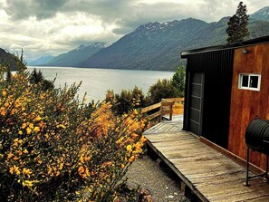 A cabin with a wooden deck overlooking a lake, surrounded by mountains and autumn foliage. - Ideal apartment for a couple with views of the lake and the hills in Villa la Angostura (Villa la Angostura)