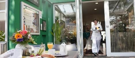 A hotel entrance with a green exterior, a table set with breakfast, and a person in uniform.