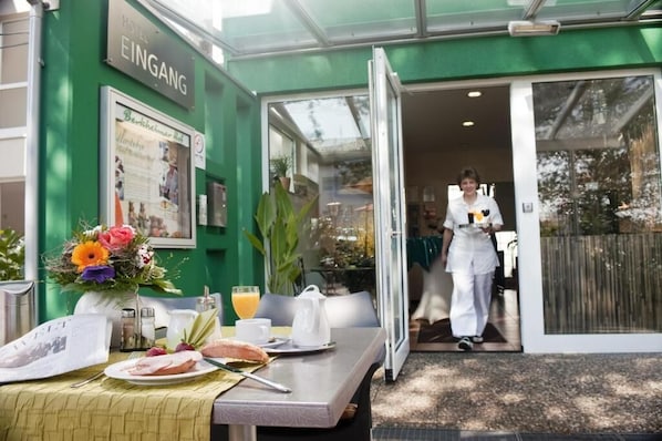 Entrada de un hotel con fachada verde, una mesa dispuesta para el desayuno y una persona con uniforme.