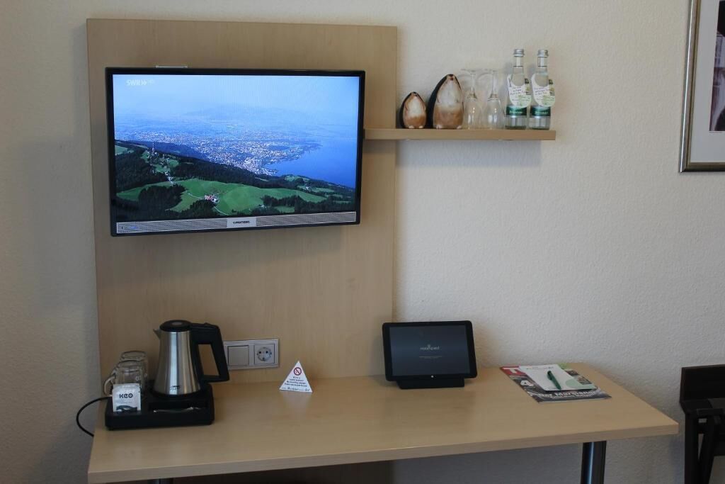 Ein Hotelrezeptionstresen mit einem Flachbildfernseher, der eine Landschaft zeigt, einer Kaffeemaschine, einem Tablet und einem Regal mit Flaschen.