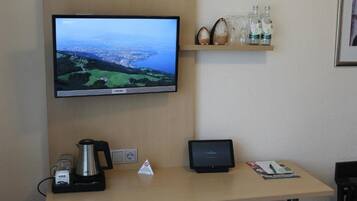 Ein Hotelrezeptionstresen mit einem Flachbildfernseher, der eine Landschaft zeigt, einer Kaffeemaschine, einem Tablet und einem Regal mit Flaschen.